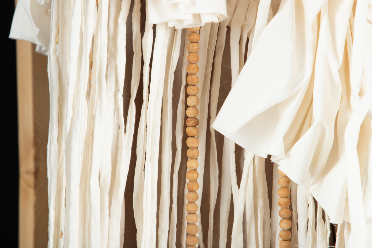 Tekstil skulptur i lyse farger som henger fra en frittstående firesidet ramme lagd av stenderverk i tre.