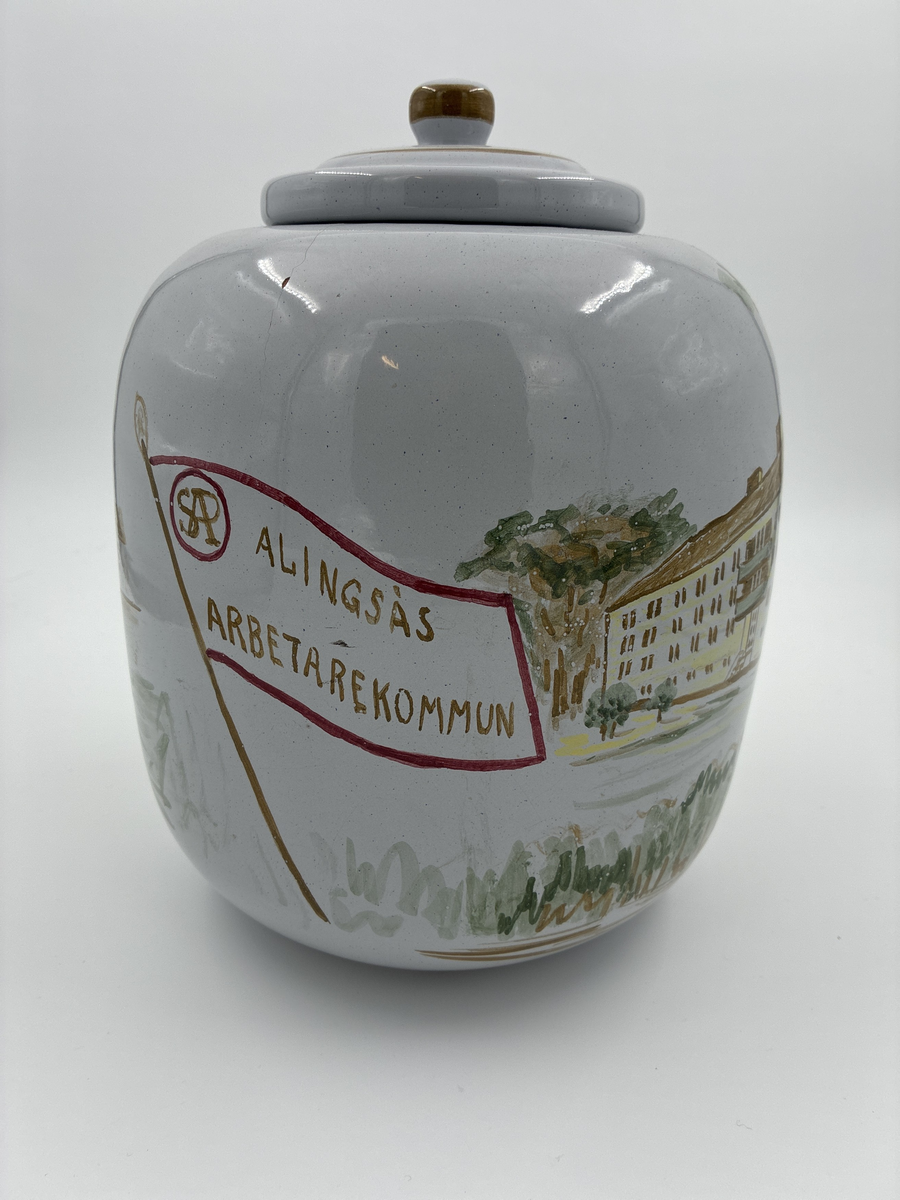 Urna med lock. Vitglaserad med handmålat motiv föreställande Alingsås fattigstuga, fana för Alingsås arbetarekommun och ålderdomshem.