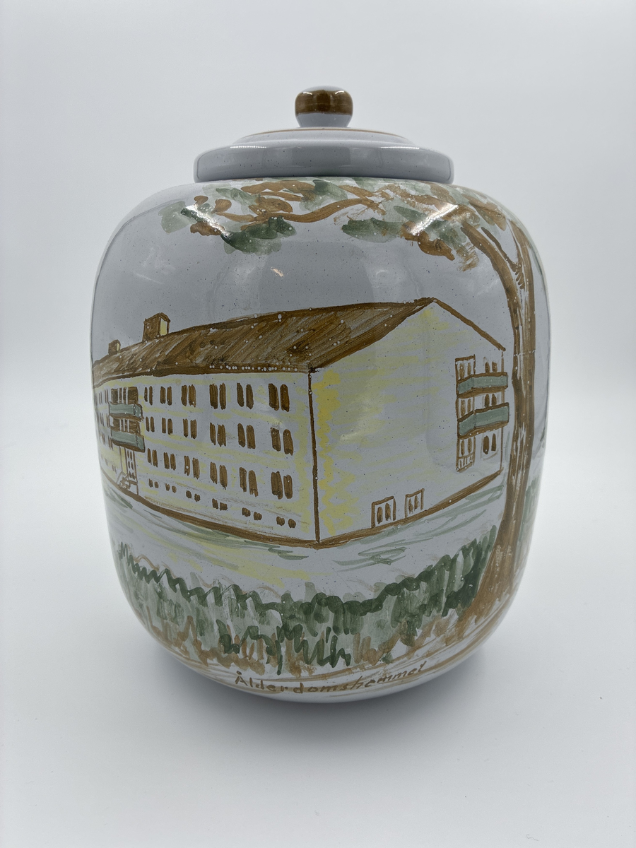 Urna med lock. Vitglaserad med handmålat motiv föreställande Alingsås fattigstuga, fana för Alingsås arbetarekommun och ålderdomshem.