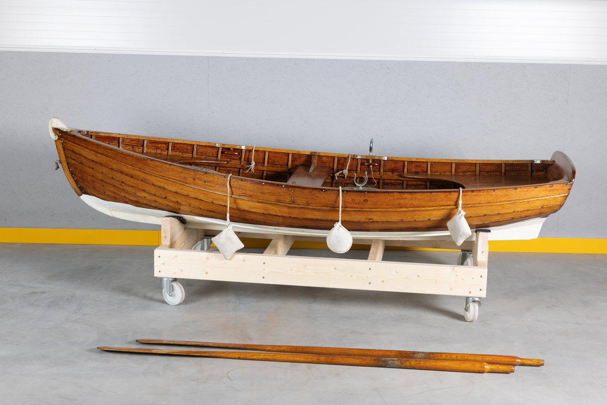 Kutterjolle med tiljer og to årer. Åregafler i metall og stoppede fendere i tekstil. Kjølen og nedre del av skroget er malt hvit.