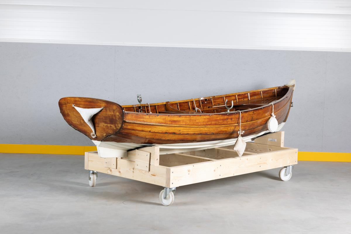 Kutterjolle med tiljer og to årer. Åregafler i metall og stoppede fendere i tekstil. Kjølen og nedre del av skroget er malt hvit.