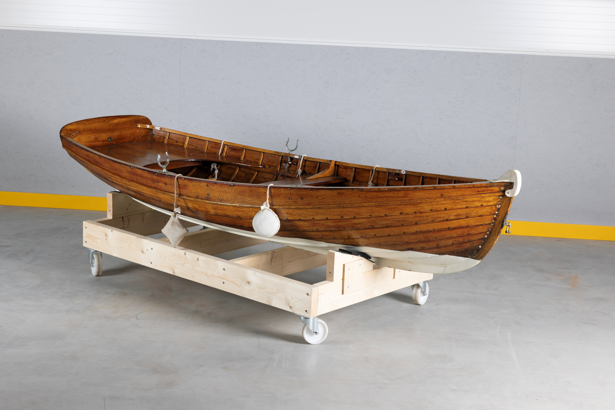 Kutterjolle med tiljer og to årer. Åregafler i metall og stoppede fendere i tekstil. Kjølen og nedre del av skroget er malt hvit.