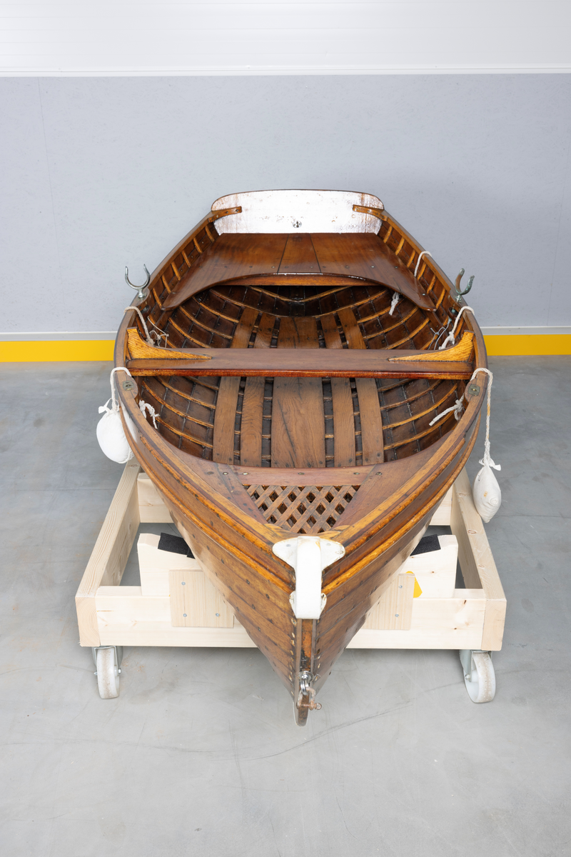 Kutterjolle med tiljer og to årer. Åregafler i metall og stoppede fendere i tekstil. Kjølen og nedre del av skroget er malt hvit.