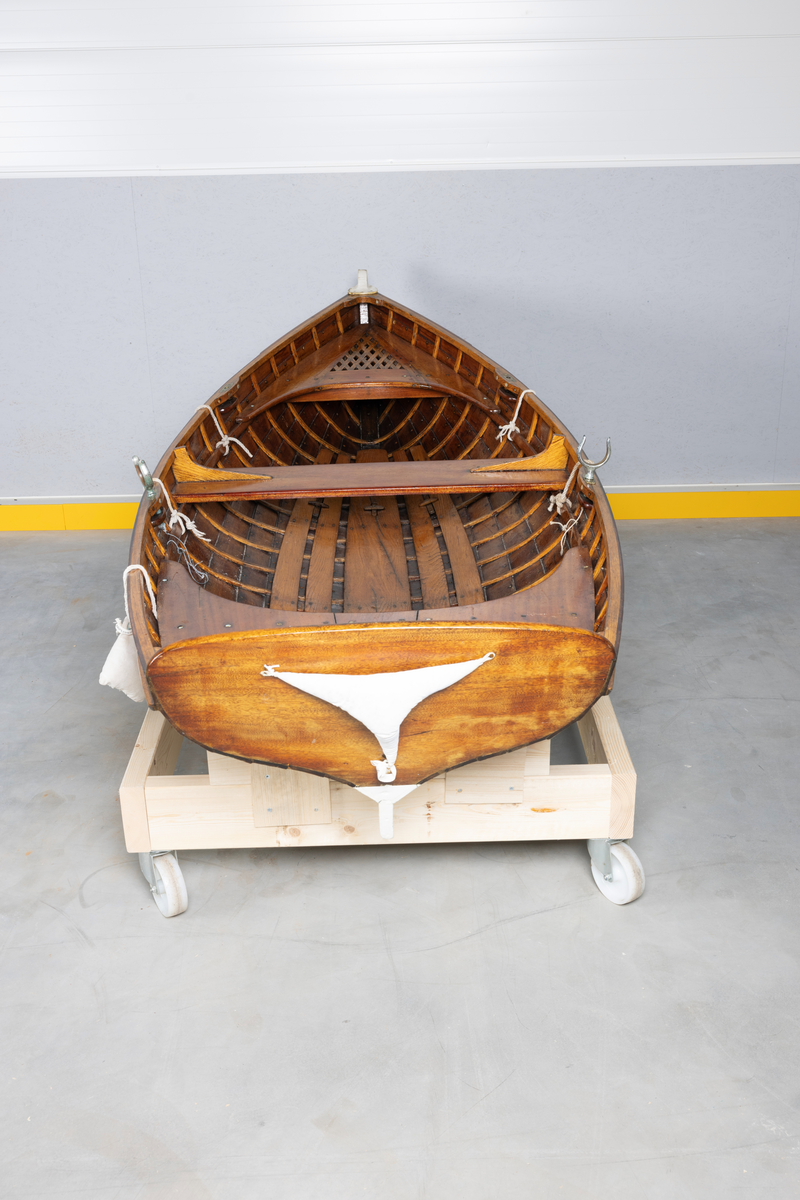 Kutterjolle med tiljer og to årer. Åregafler i metall og stoppede fendere i tekstil. Kjølen og nedre del av skroget er malt hvit.