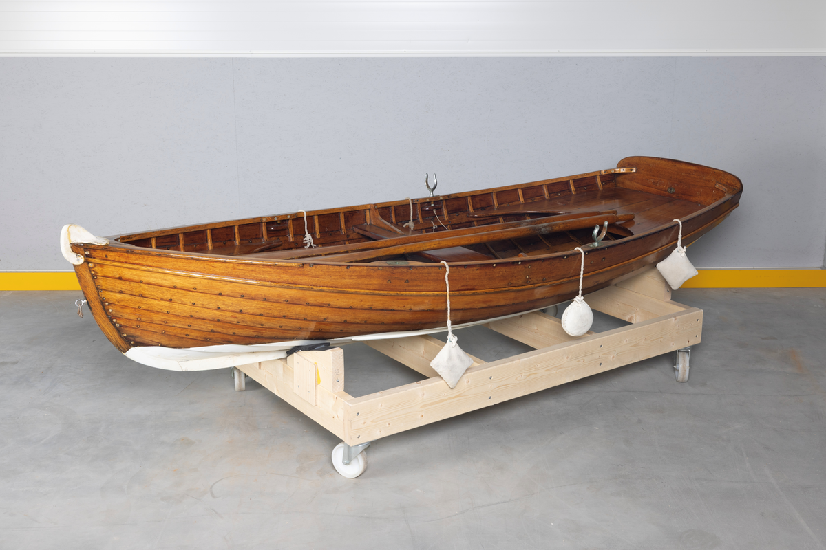 Kutterjolle med tiljer og to årer. Åregafler i metall og stoppede fendere i tekstil. Kjølen og nedre del av skroget er malt hvit.