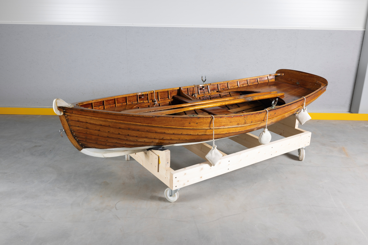 Kutterjolle med tiljer og to årer. Åregafler i metall og stoppede fendere i tekstil. Kjølen og nedre del av skroget er malt hvit.