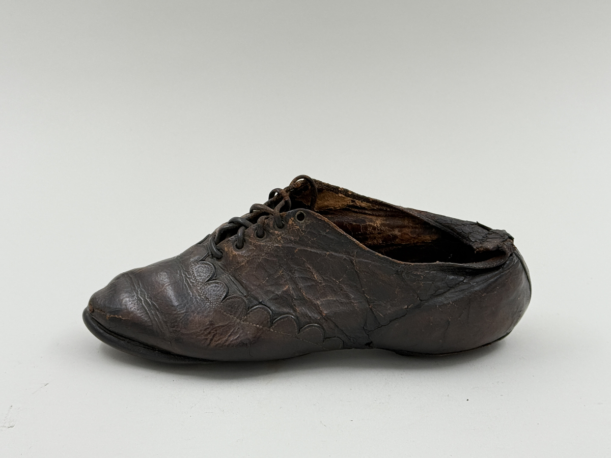 Damsko gjord på rak läst, spetsig lätt rundad tå. Lädret över tån skuret med dekorativt mönster av halvcirklar. Fem par snörhål, vissa med mässingsöljetter. Lädersula, liten klack. Lädret i hälen skadat.
 