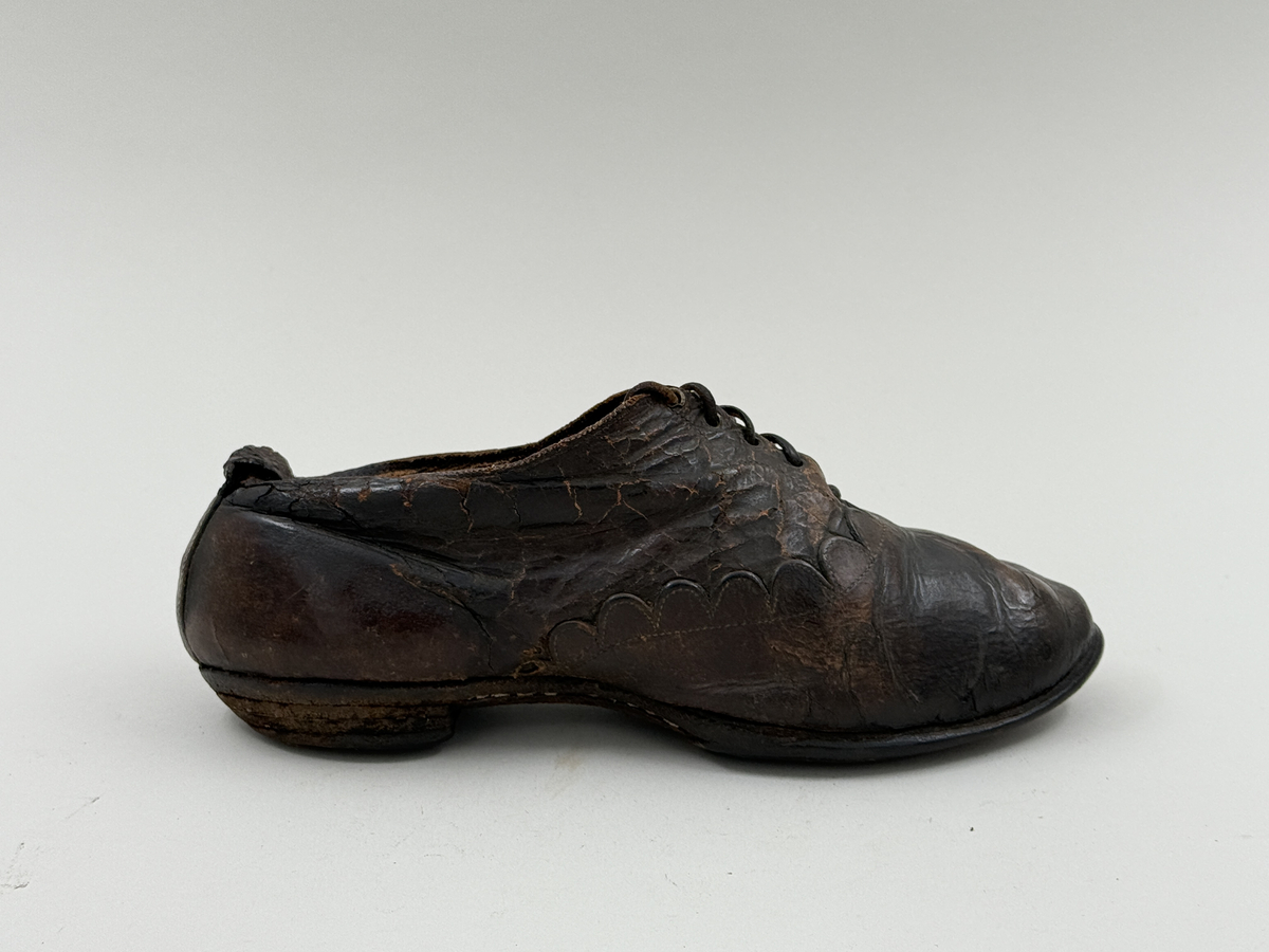 Damsko gjord på rak läst, spetsig lätt rundad tå. Lädret över tån skuret med dekorativt mönster av halvcirklar. Fem par snörhål, vissa med mässingsöljetter. Lädersula, liten klack. Lädret i hälen skadat.
 