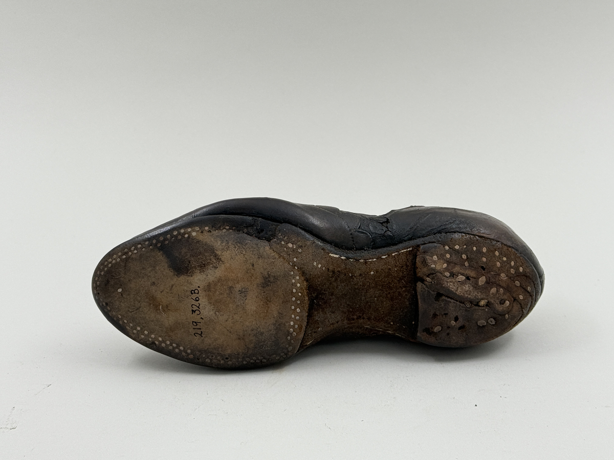 Damsko gjord på rak läst, spetsig lätt rundad tå. Lädret över tån skuret med dekorativt mönster av halvcirklar. Fem par snörhål, vissa med mässingsöljetter. Lädersula, liten klack. Lädret i hälen skadat.
 
