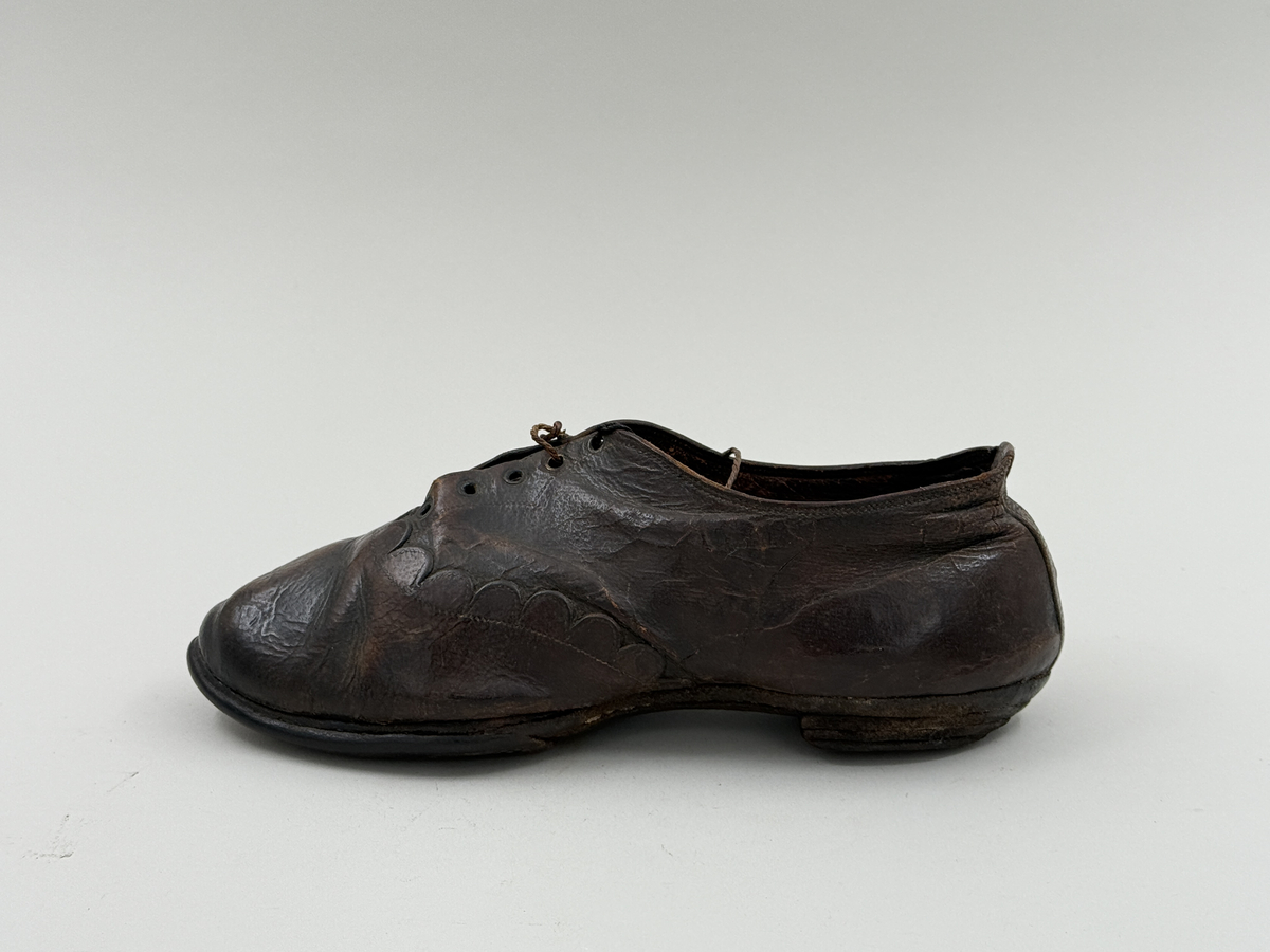 Damsko gjord på rak läst, spetsig lätt rundad tå. Lädret över tån skuret med dekorativt mönster av halvcirklar. Fem par snörhål, vissa med mässingsöljetter. Lädersula, liten klack. 
 