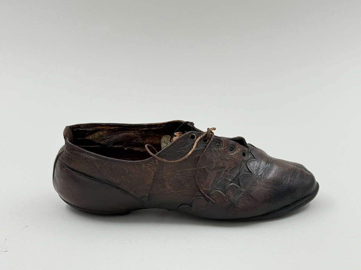 Damsko gjord på rak läst, spetsig lätt rundad tå. Lädret över tån skuret med dekorativt mönster av halvcirklar. Fem par snörhål, vissa med mässingsöljetter. Lädersula, liten klack. 
 