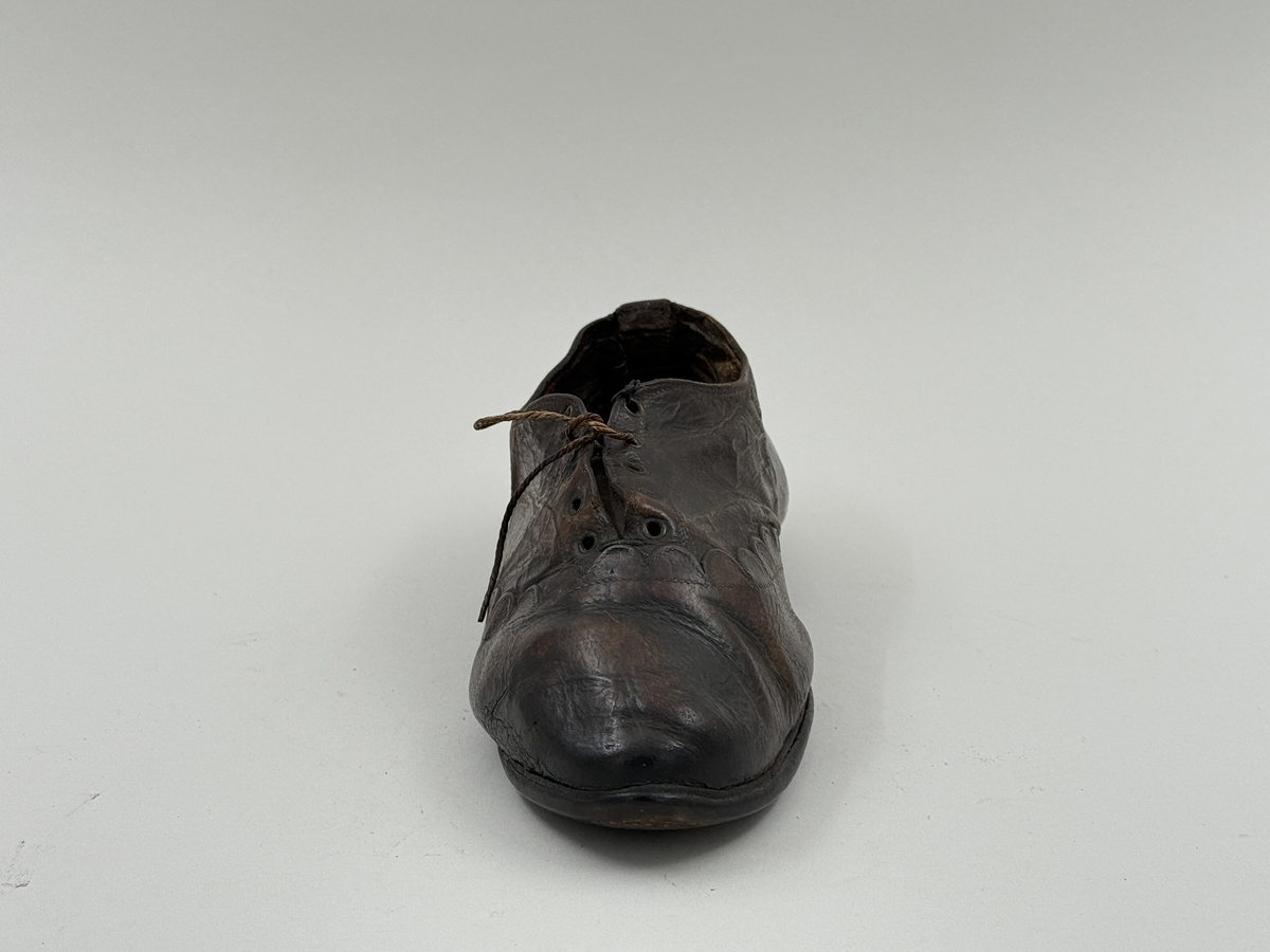 Damsko gjord på rak läst, spetsig lätt rundad tå. Lädret över tån skuret med dekorativt mönster av halvcirklar. Fem par snörhål, vissa med mässingsöljetter. Lädersula, liten klack. 
 