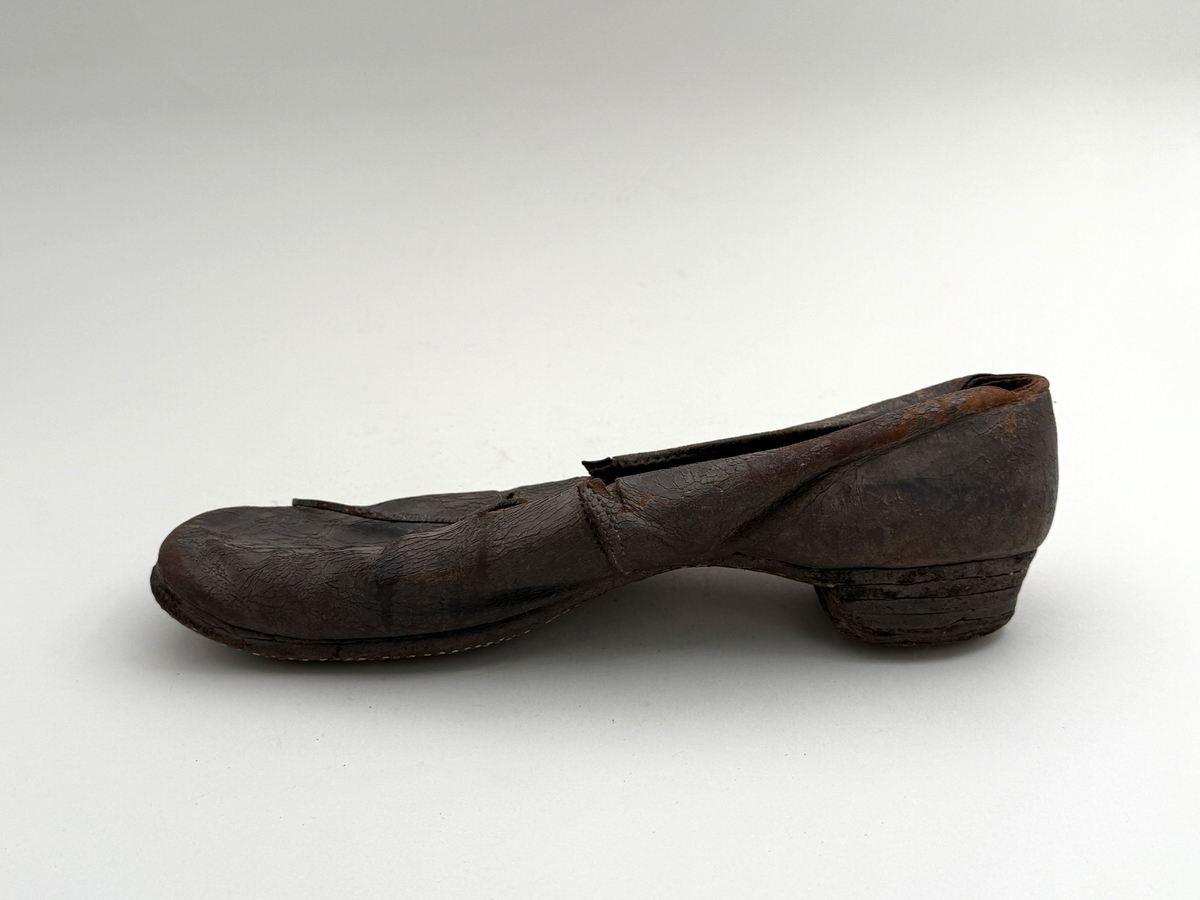 Sko för högerfot av svart läder, träpliggad sula av läder samt med becksömnad. Lädret löper över i två sleifer över plösen, genom plösen och sleifarna hål för snöre. Lädret skadat vid sleifarna. 