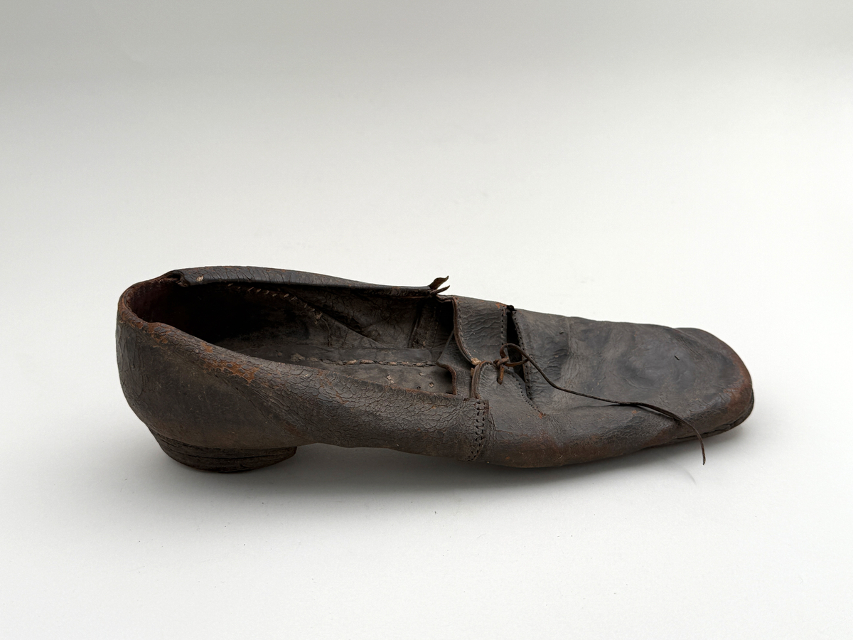 Sko för högerfot av svart läder, träpliggad sula av läder samt med becksömnad. Lädret löper över i två sleifer över plösen, genom plösen och sleifarna hål för snöre. Lädret skadat vid sleifarna. 