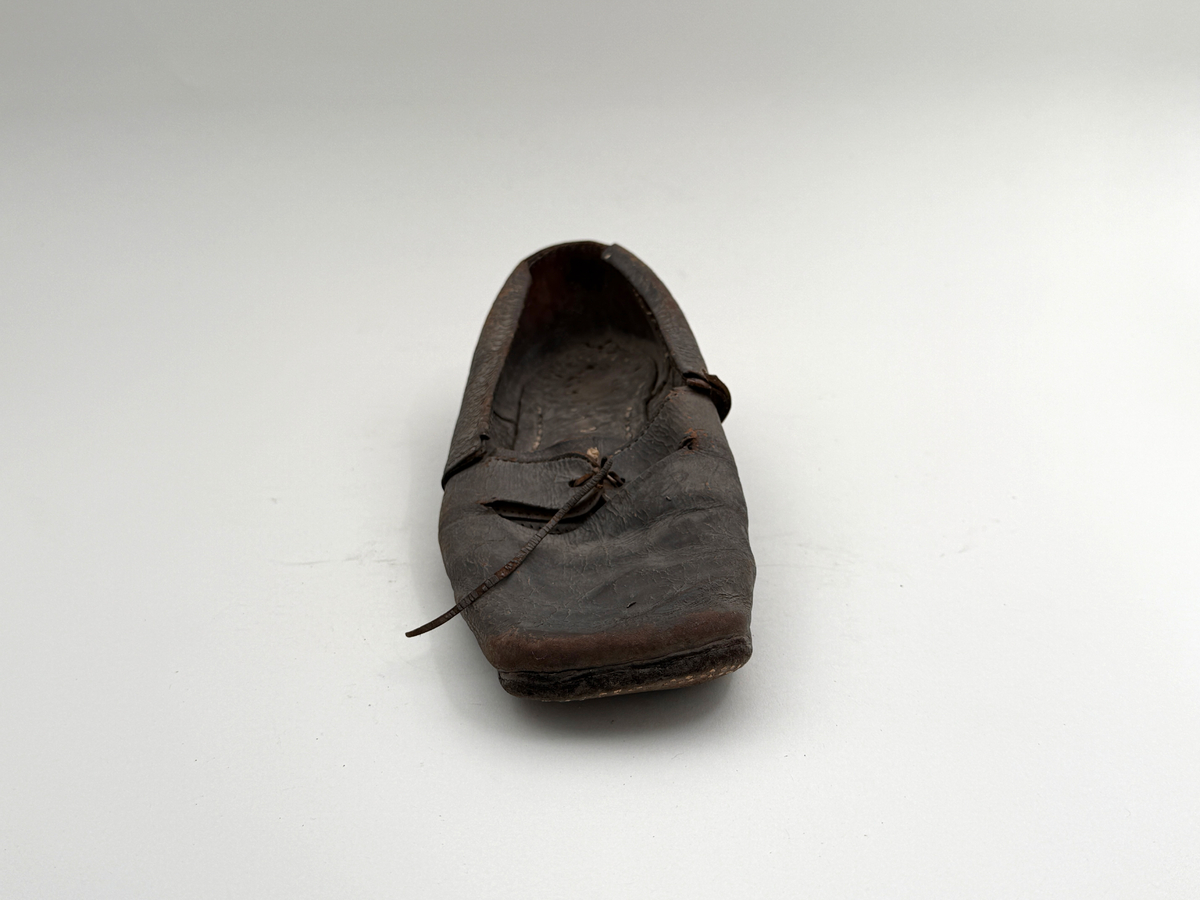 Sko för högerfot av svart läder, träpliggad sula av läder samt med becksömnad. Lädret löper över i två sleifer över plösen, genom plösen och sleifarna hål för snöre. Lädret skadat vid sleifarna. 