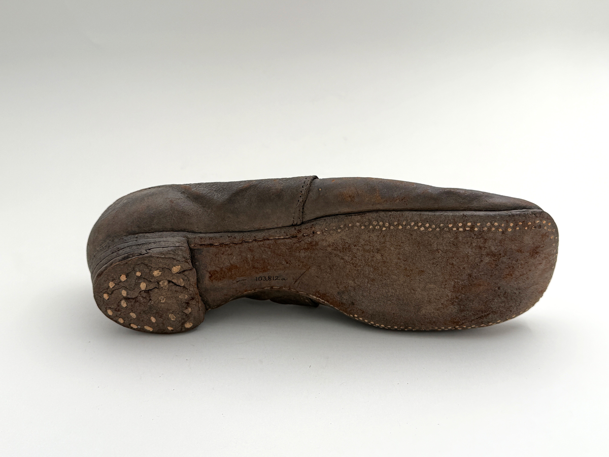 Sko för högerfot av svart läder, träpliggad sula av läder samt med becksömnad. Lädret löper över i två sleifer över plösen, genom plösen och sleifarna hål för snöre. Lädret skadat vid sleifarna. 