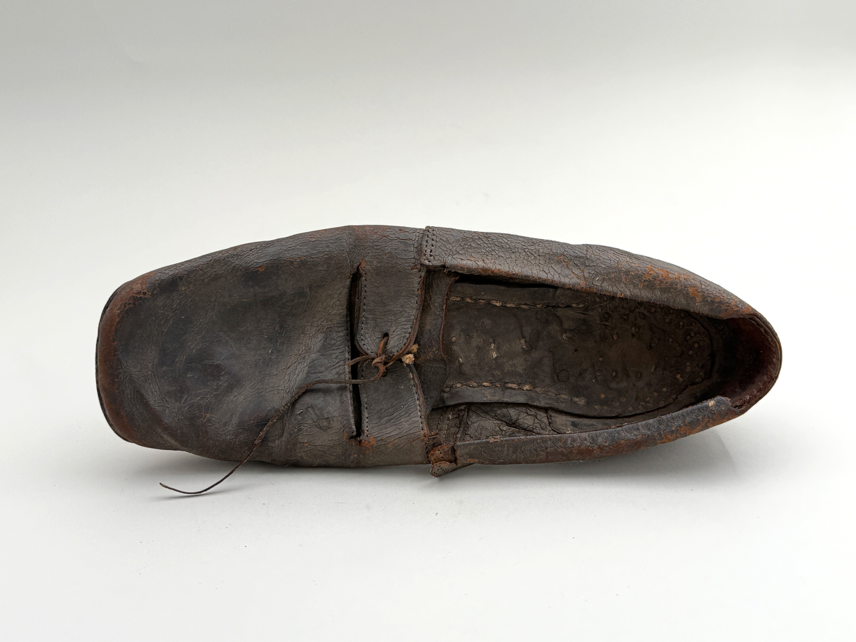 Sko för högerfot av svart läder, träpliggad sula av läder samt med becksömnad. Lädret löper över i två sleifer över plösen, genom plösen och sleifarna hål för snöre. Lädret skadat vid sleifarna. 