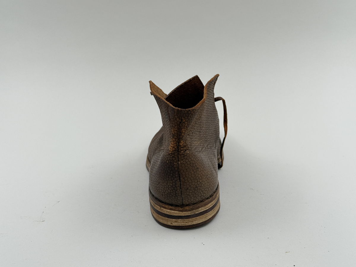 Låg snörkängor för vänster fot, av mörkbrunt läder med präglat stjärnmönster. Kanten upptill rakt avskuren. 4 par snörhål. Slitsula av läder med mellansula av näver, becksömmade. Innersula av näver. Klack järnpliggad. Nygjord efter gammal modell av givaren för museet.