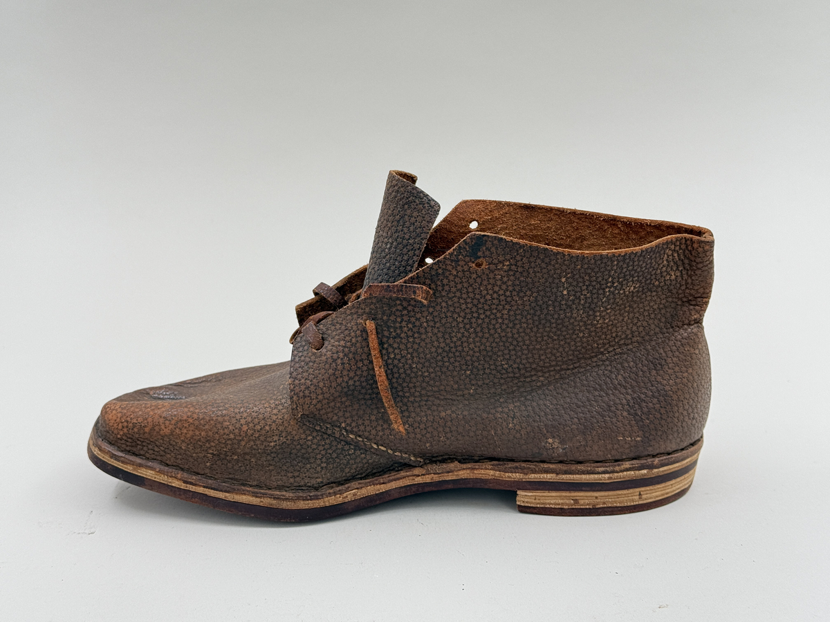 Låg snörkängor för höger fot,  av mörkbrunt läder med präglat stjärnmönster. Kanten upptill rakt avskuren. 4 par snörhål. Slitsula av läder med mellansula av näver, becksömmade. Innersula av näver. Klack järnpliggad. Nygjord efter gammal modell av givaren för museet.