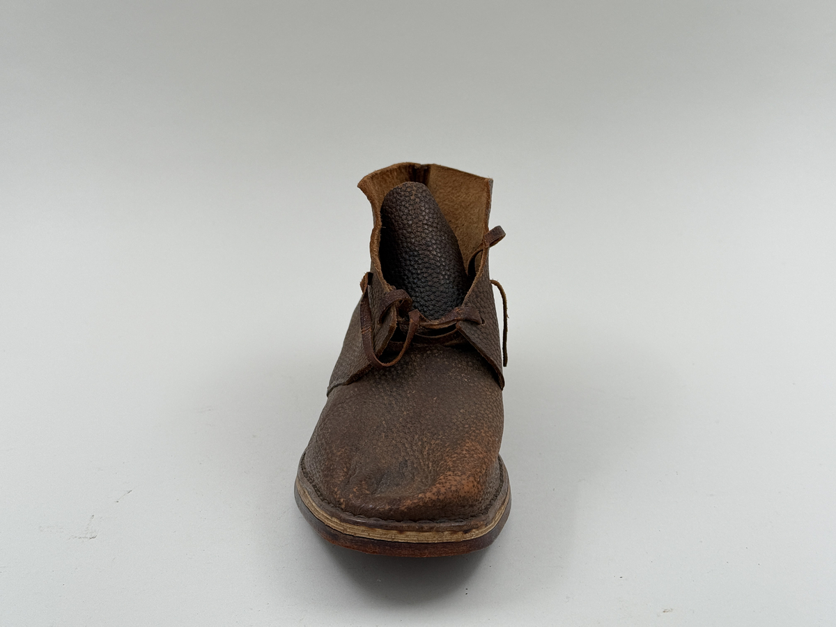 Låg snörkängor för höger fot,  av mörkbrunt läder med präglat stjärnmönster. Kanten upptill rakt avskuren. 4 par snörhål. Slitsula av läder med mellansula av näver, becksömmade. Innersula av näver. Klack järnpliggad. Nygjord efter gammal modell av givaren för museet.