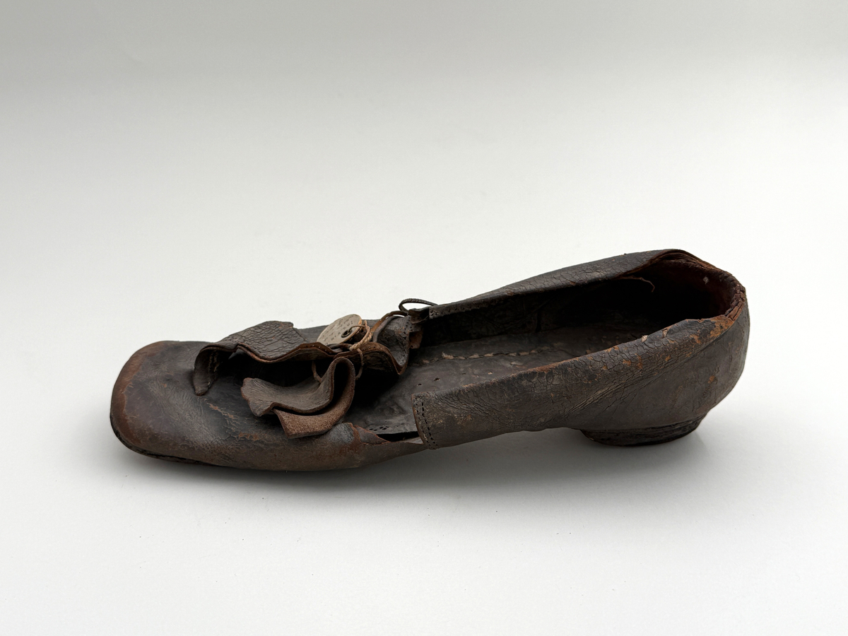 Sko för vänsterfot av svart läder, träpliggad sula av läder samt med becksömnad. Lädret löper över i två sleifer över plösen, genom plösen och sleifarna hål för snöre.
 