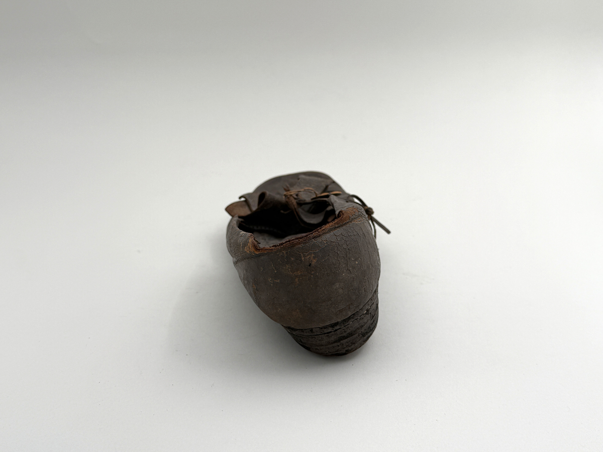 Sko för vänsterfot av svart läder, träpliggad sula av läder samt med becksömnad. Lädret löper över i två sleifer över plösen, genom plösen och sleifarna hål för snöre.
 