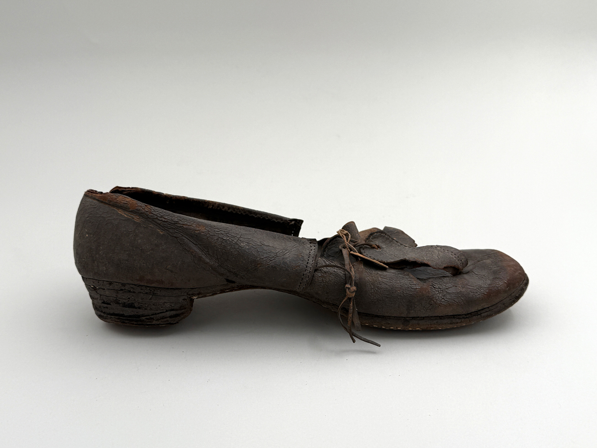 Sko för vänsterfot av svart läder, träpliggad sula av läder samt med becksömnad. Lädret löper över i två sleifer över plösen, genom plösen och sleifarna hål för snöre.
 