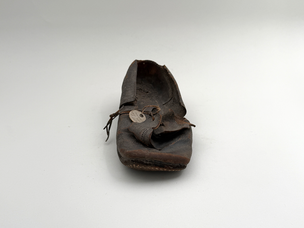 Sko för vänsterfot av svart läder, träpliggad sula av läder samt med becksömnad. Lädret löper över i två sleifer över plösen, genom plösen och sleifarna hål för snöre.
 