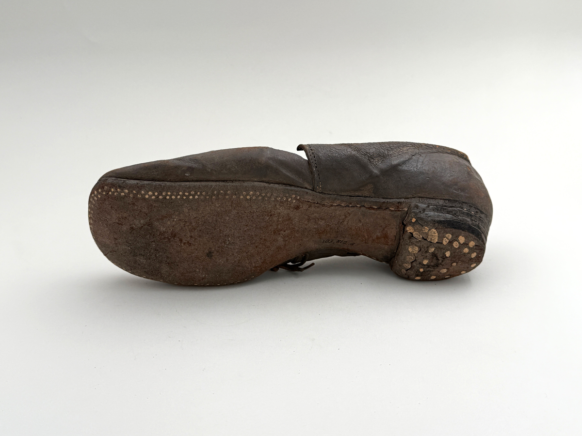 Sko för vänsterfot av svart läder, träpliggad sula av läder samt med becksömnad. Lädret löper över i två sleifer över plösen, genom plösen och sleifarna hål för snöre.
 
