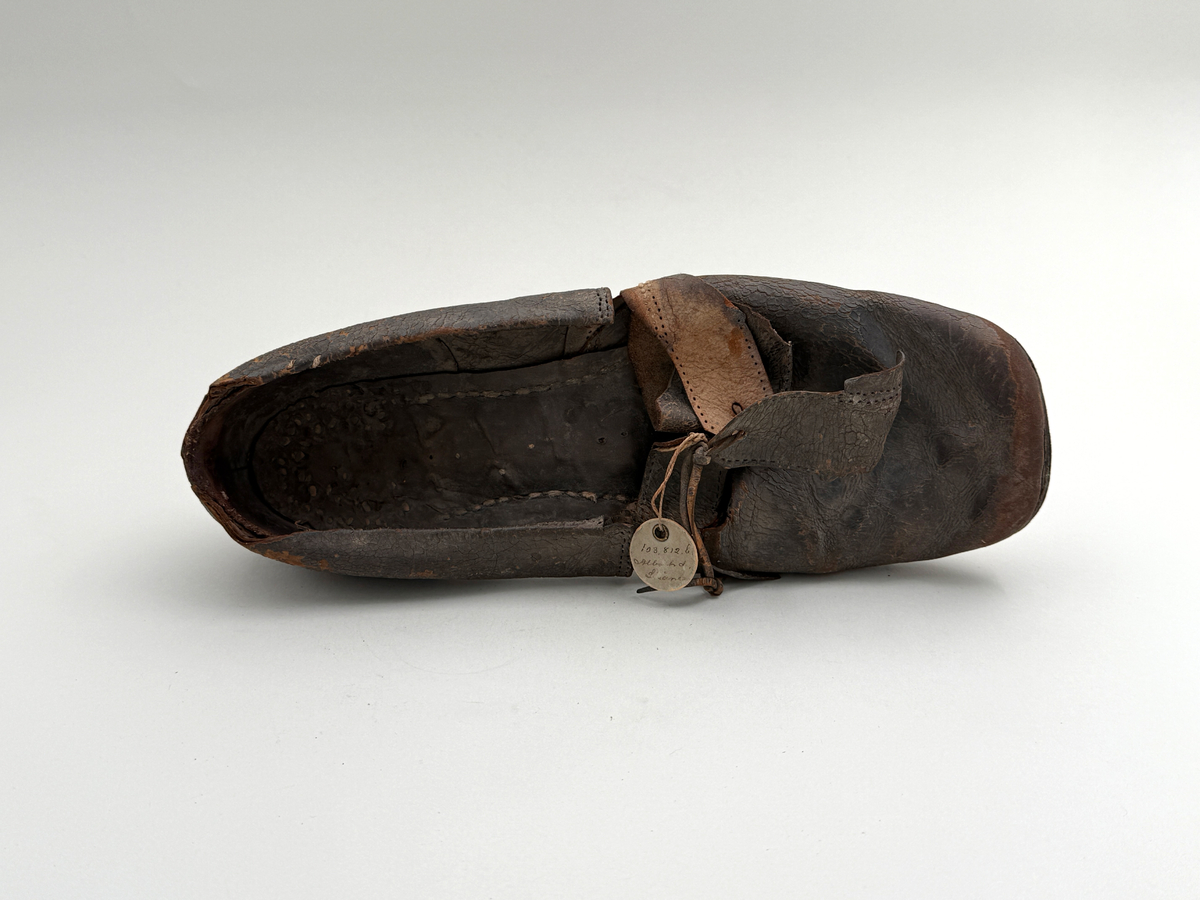 Sko för vänsterfot av svart läder, träpliggad sula av läder samt med becksömnad. Lädret löper över i två sleifer över plösen, genom plösen och sleifarna hål för snöre.
 