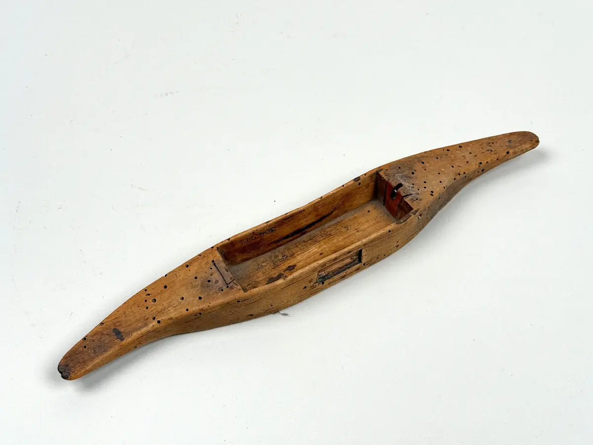 Skyttel av snidat och delvis urgröpt björk, avlång form med avsmalnande ändar. Rektangulärt uttag mitt på ena sidan för att hålla tråden, litet rektangulärt uttag på sidan. 

Metallpinne att trä trådspole på saknas. 