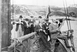 Gruppebilde av åtte tørrfiskspisende menn i søndagsklær på a