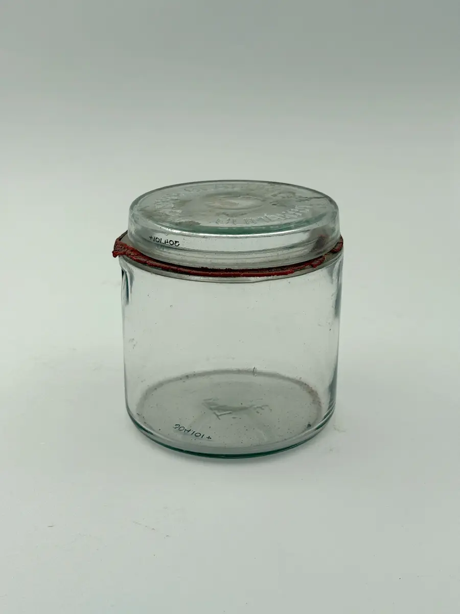 Cylindrisk burk av klarglas med lock och gummiring. Glaset formgjutet. Burken med intag i glaset på vardera sida för fästa av metallbygel som ska hålla locket på plats. Locket märkt GLIMMA GLASBRUK GLIMÅKRA, och botten märkt 3/4 LIT. SURTE. 

Metallbygel saknas. Locket sitter fast i den torkade gummiringen. 