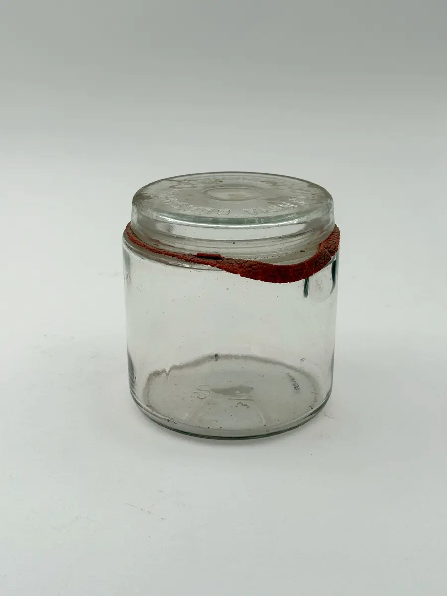 Cylindrisk burk av klarglas med lock och gummiring. Glaset formgjutet. Burken med intag i glaset på vardera sida för fästa av metallbygel som ska hålla locket på plats. Locket märkt GLIMMA GLASBRUK GLIMÅKRA, och botten märkt 3/4 LIT. SURTE. 

Metallbygel saknas. Locket sitter fast i den torkade gummiringen. 