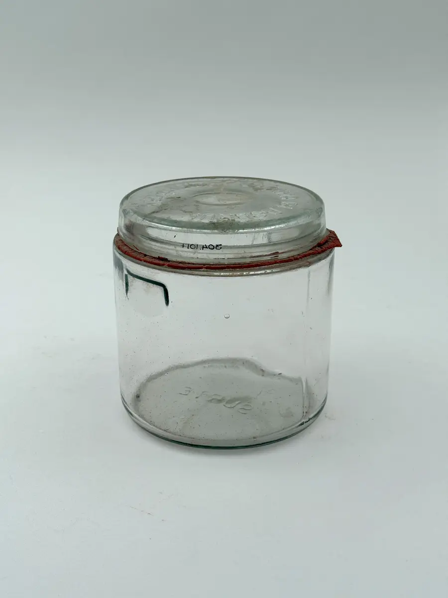 Cylindrisk burk av klarglas med lock och gummiring. Glaset formgjutet. Burken med intag i glaset på vardera sida för fästa av metallbygel som ska hålla locket på plats. Locket märkt GLIMMA GLASBRUK GLIMÅKRA, och botten märkt 3/4 LIT. SURTE. 

Metallbygel saknas. Locket sitter fast i den torkade gummiringen. 