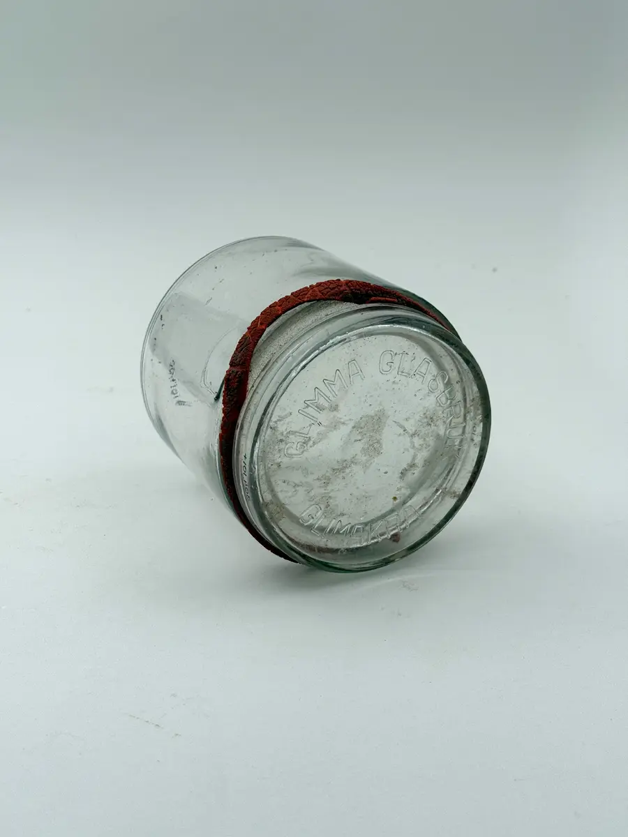 Cylindrisk burk av klarglas med lock och gummiring. Glaset formgjutet. Burken med intag i glaset på vardera sida för fästa av metallbygel som ska hålla locket på plats. Locket märkt GLIMMA GLASBRUK GLIMÅKRA, och botten märkt 3/4 LIT. SURTE. 

Metallbygel saknas. Locket sitter fast i den torkade gummiringen. 