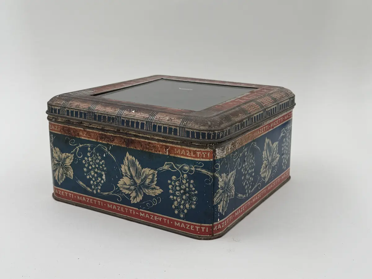 Kvadratisk burk av plåt med lock, locket med infällt glas. Tryckt dekor av vinrankor och vindruvor mot blå bakgrund, Smala röda bårder upptill och nedtill med texten MAZETTI.