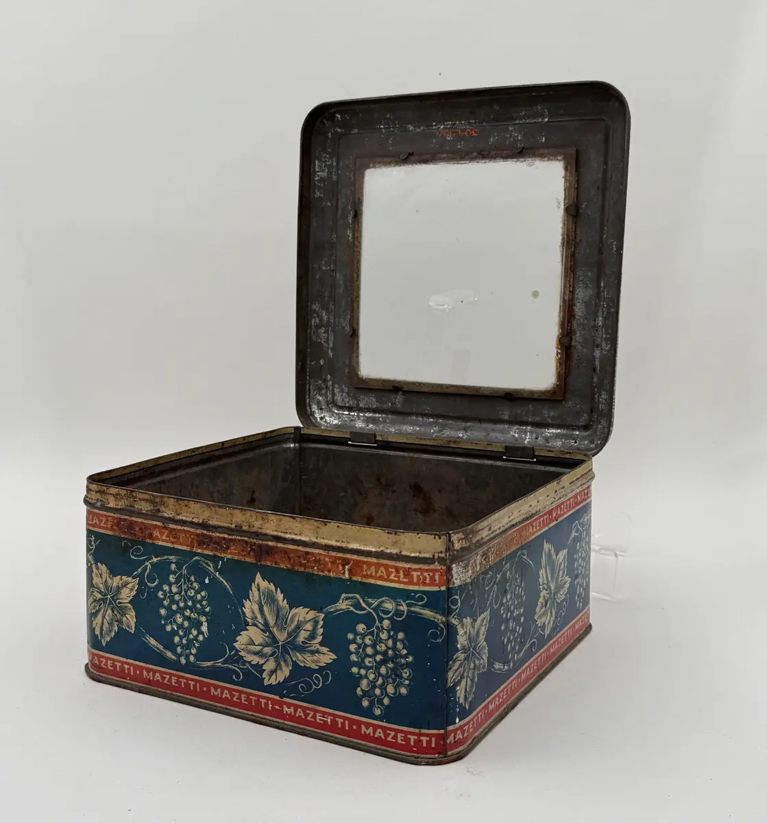 Kvadratisk burk av plåt med lock, locket med infällt glas. Tryckt dekor av vinrankor och vindruvor mot blå bakgrund, Smala röda bårder upptill och nedtill med texten MAZETTI.