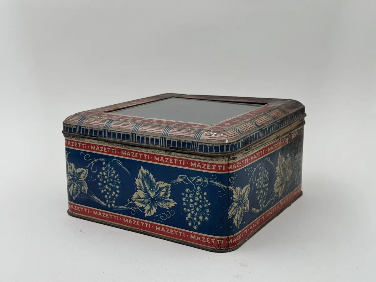 Kvadratisk burk av plåt med lock, locket med infällt glas. Tryckt dekor av vinrankor och vindruvor mot blå bakgrund, Smala röda bårder upptill och nedtill med texten MAZETTI.
