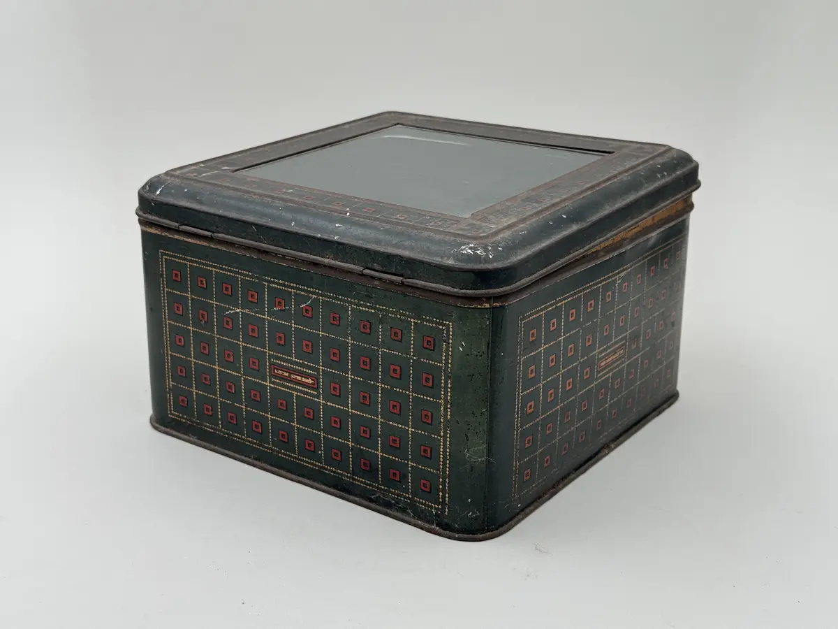 Kvadratisk burk med lock, locket med infällt glas. Målad i mörkgrönt med rutig dekor i guld och rött. Undersidan med firmamärke i svart: Carl P. Påhlssons Karamell- och konfektfabrik Malmö. 