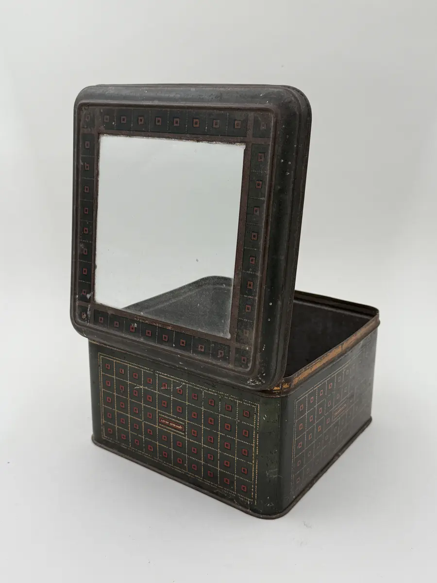 Kvadratisk burk med lock, locket med infällt glas. Målad i mörkgrönt med rutig dekor i guld och rött. Undersidan med firmamärke i svart: Carl P. Påhlssons Karamell- och konfektfabrik Malmö. 