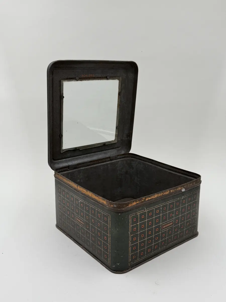 Kvadratisk burk med lock, locket med infällt glas. Målad i mörkgrönt med rutig dekor i guld och rött. Undersidan med firmamärke i svart: Carl P. Påhlssons Karamell- och konfektfabrik Malmö. 