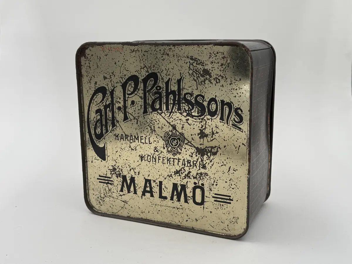 Kvadratisk burk med lock, locket med infällt glas. Målad i mörkgrönt med rutig dekor i guld och rött. Undersidan med firmamärke i svart: Carl P. Påhlssons Karamell- och konfektfabrik Malmö. 