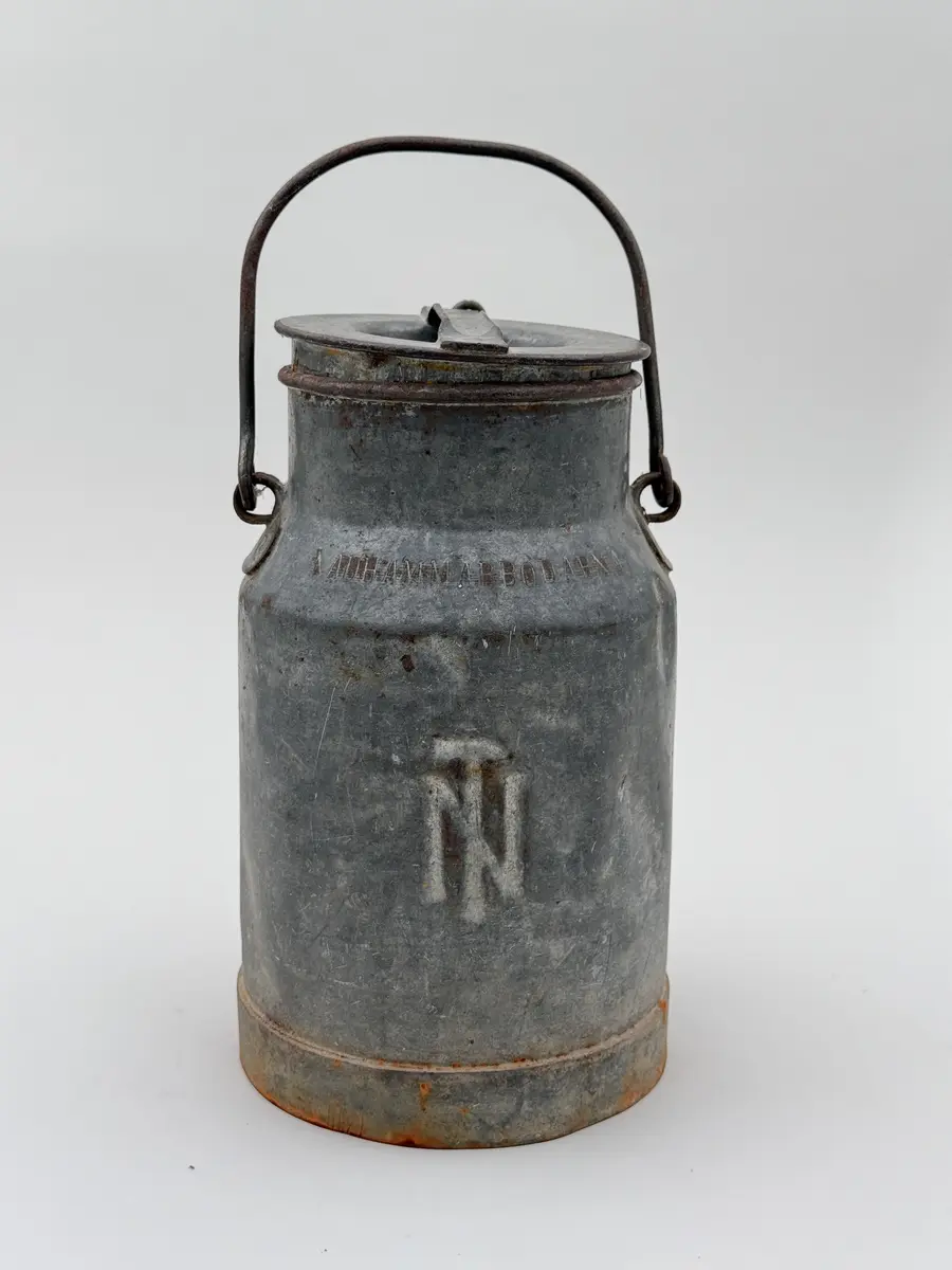 Cylindrisk mjölkflaska av plåt med något indragen hals. Platt lock samt uppsvängt handtag fäst i metallöglor på vardera sida. Märkt NÅDHAMMARBODARNA i låg relief samt med ett N med en hammare igenom i relief.

Locket fastkilat.