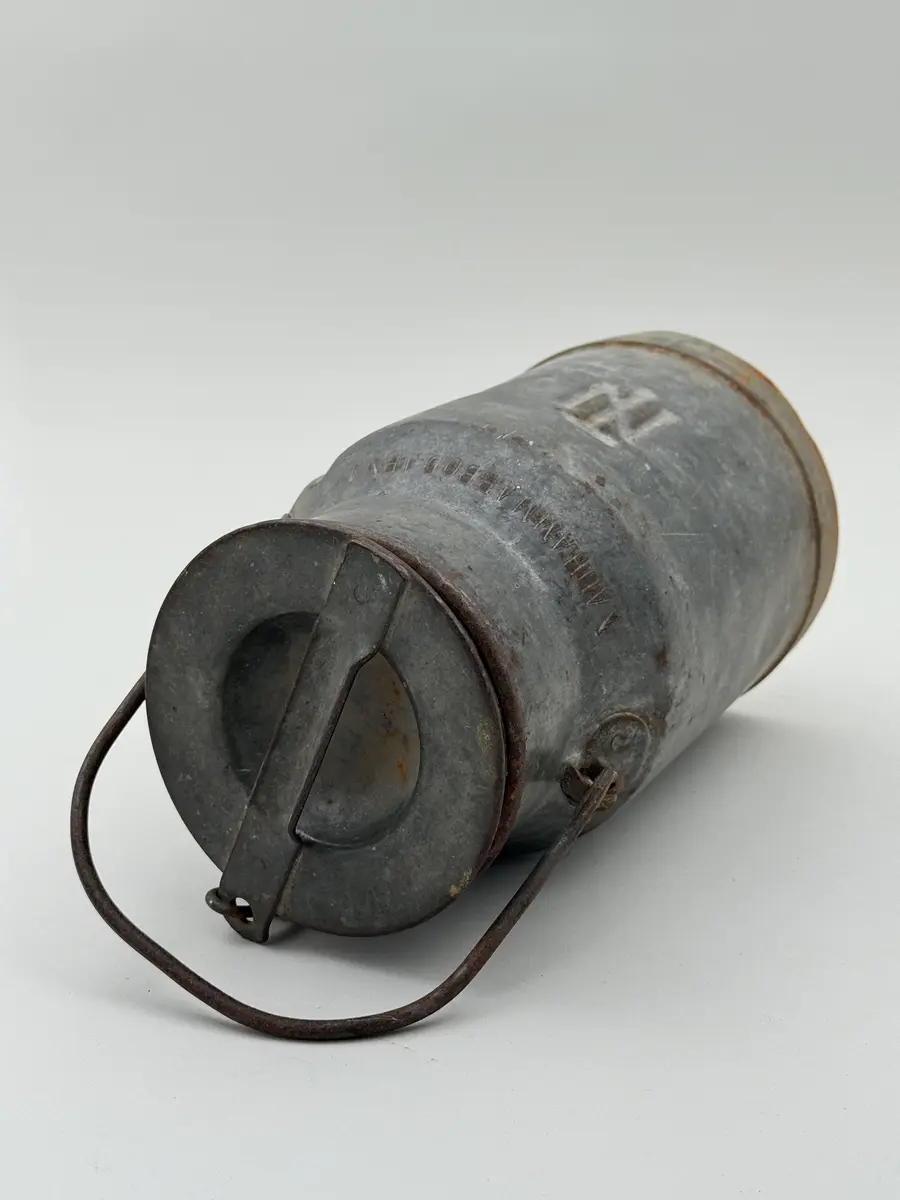 Cylindrisk mjölkflaska av plåt med något indragen hals. Platt lock samt uppsvängt handtag fäst i metallöglor på vardera sida. Märkt NÅDHAMMARBODARNA i låg relief samt med ett N med en hammare igenom i relief.

Locket fastkilat.