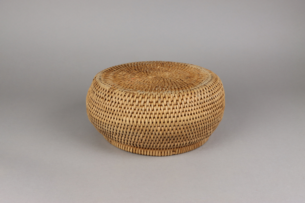 Korg bunden av kluvna rottågor över vidjor med löst lock. Rund bukig form med platt botten och platt lock.