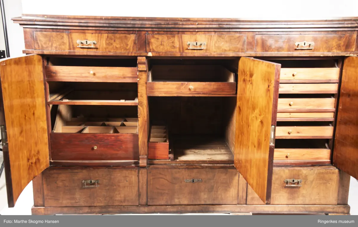 Stor seksjon i mahogni med skuffer og tre skapdører. Innenfor skapdørene er det. To av skuffene er skåret ut for hånd med plass til bestikk og kopper.