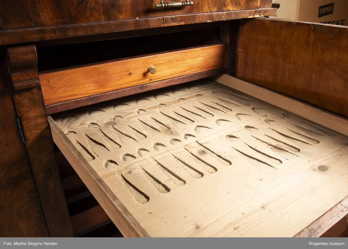 Stor seksjon i mahogni med skuffer og tre skapdører. Innenfor skapdørene er det. To av skuffene er skåret ut for hånd med plass til bestikk og kopper.