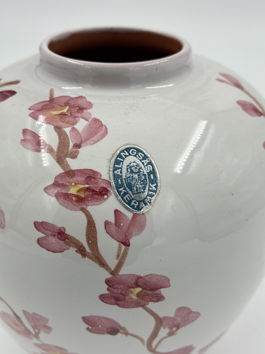 Kruka med lock. Vit glaserad, dekorerad med grenar i brunt och små rosa blommor. Även dekorerad på locket. Glaserad på insidan. På skuldran sitter en oval klisterlapp i blått och silver, Jonas Alströmer i mitten och texten Alingsås Keramik runt om.
Stämplad i botten: Aås 838 2 samt en otydlig signatur i rosa glasyr.
