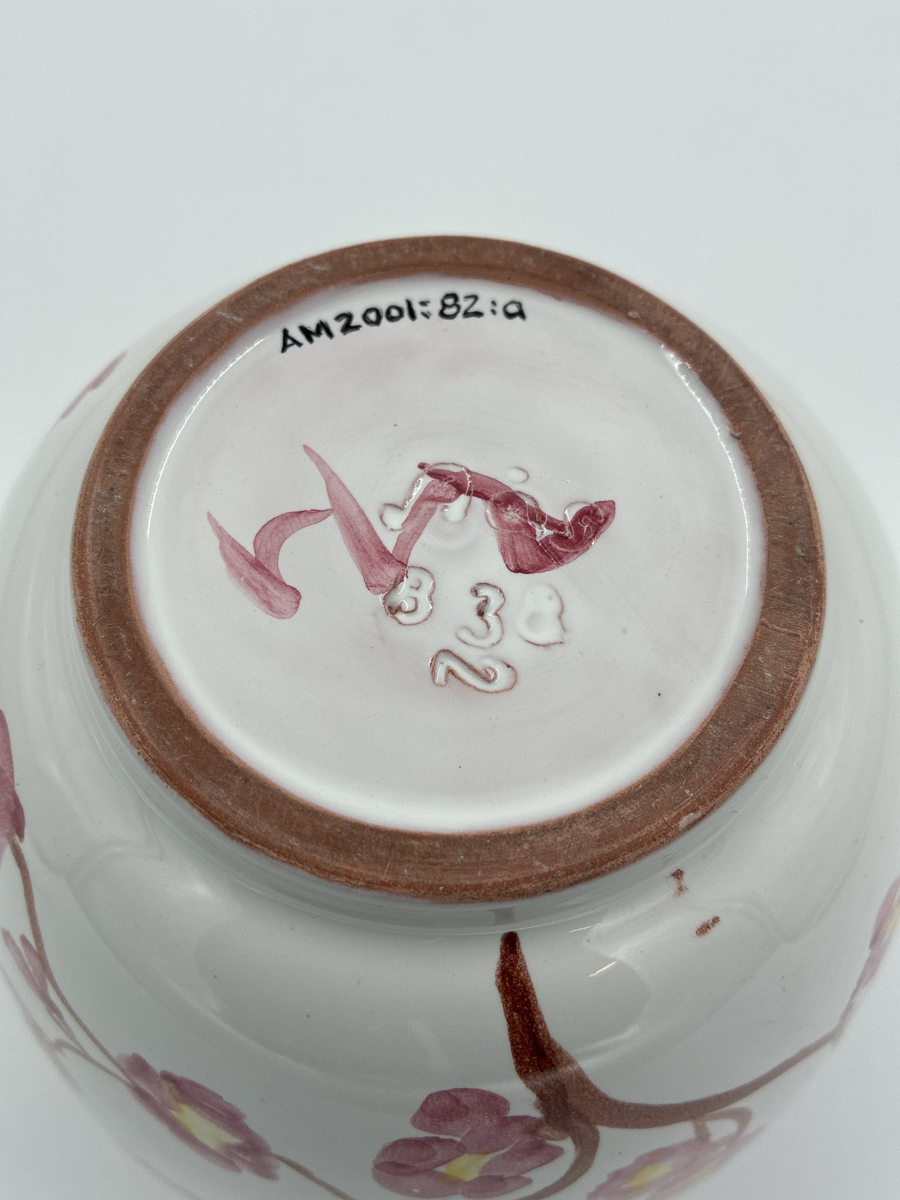 Kruka med lock. Vit glaserad, dekorerad med grenar i brunt och små rosa blommor. Även dekorerad på locket. Glaserad på insidan. På skuldran sitter en oval klisterlapp i blått och silver, Jonas Alströmer i mitten och texten Alingsås Keramik runt om.
Stämplad i botten: Aås 838 2 samt en otydlig signatur i rosa glasyr.
